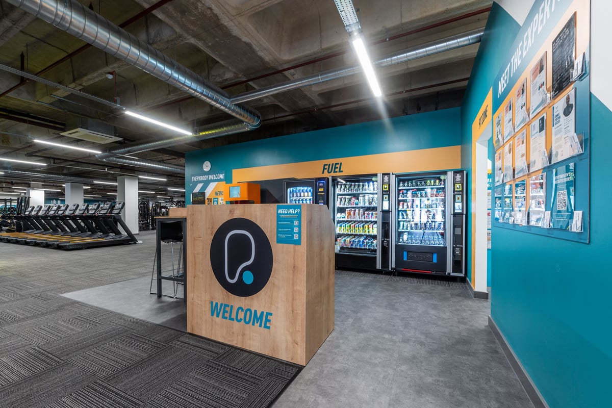 PureGym London Ealing Broadway: Rows of cardio machines, vending fridges, a wooden welcome podium, and wall graphics.