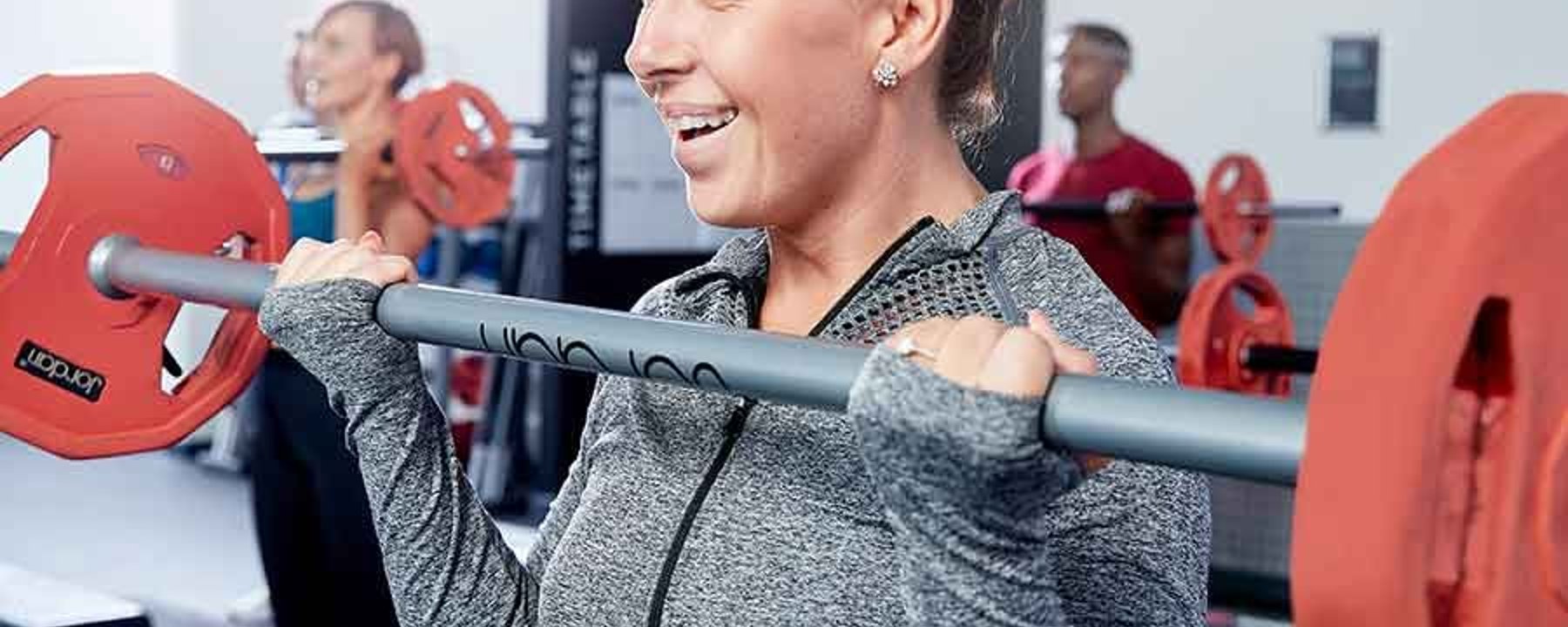 Frau hebt Langhantel im Fitnessstudio, andere trainieren im Hintergrund