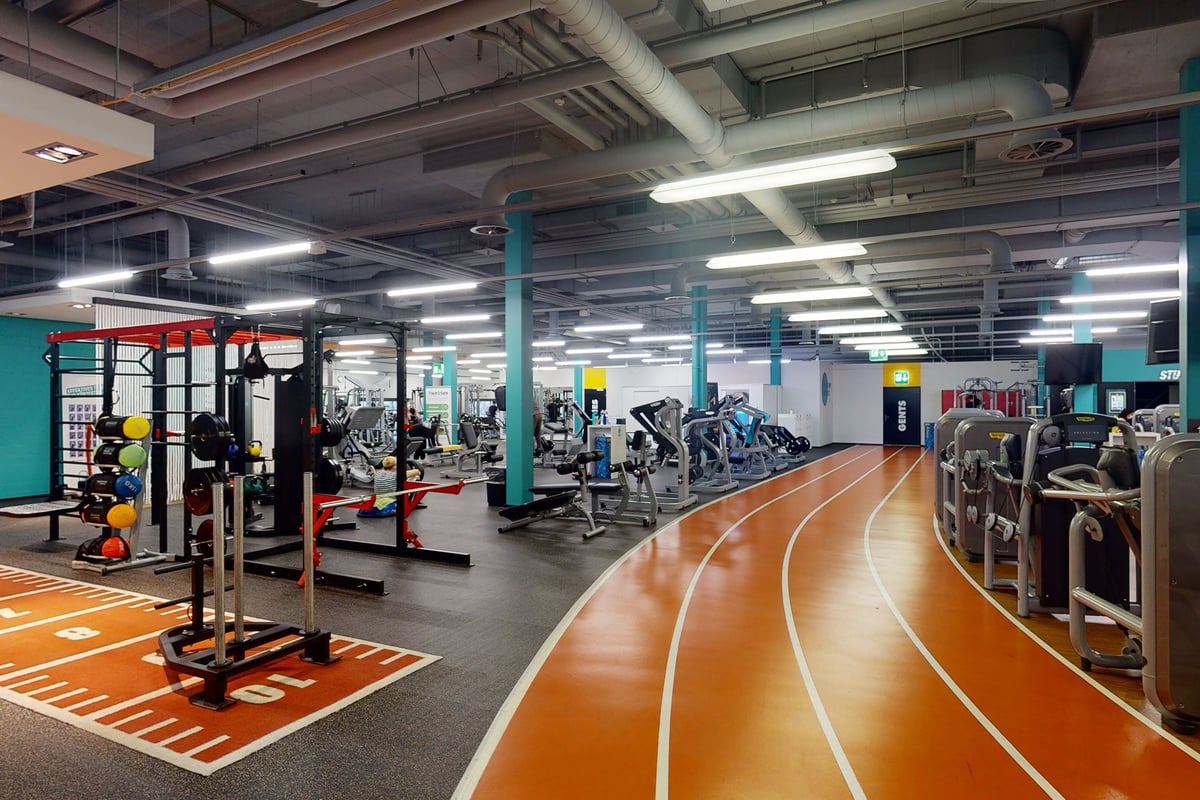 PureGym Winterthur Töss: Salle de sport avec haltères, appareils de résistance, piste caoutchoutée, plateformes de musculation et plafond apparent.