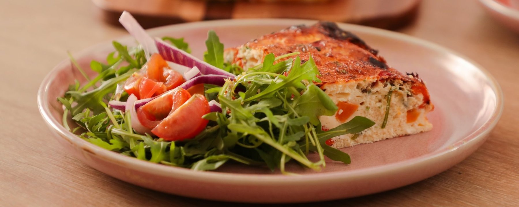 Slice of frittata with rocket salad, tomato and red onion on pink plate