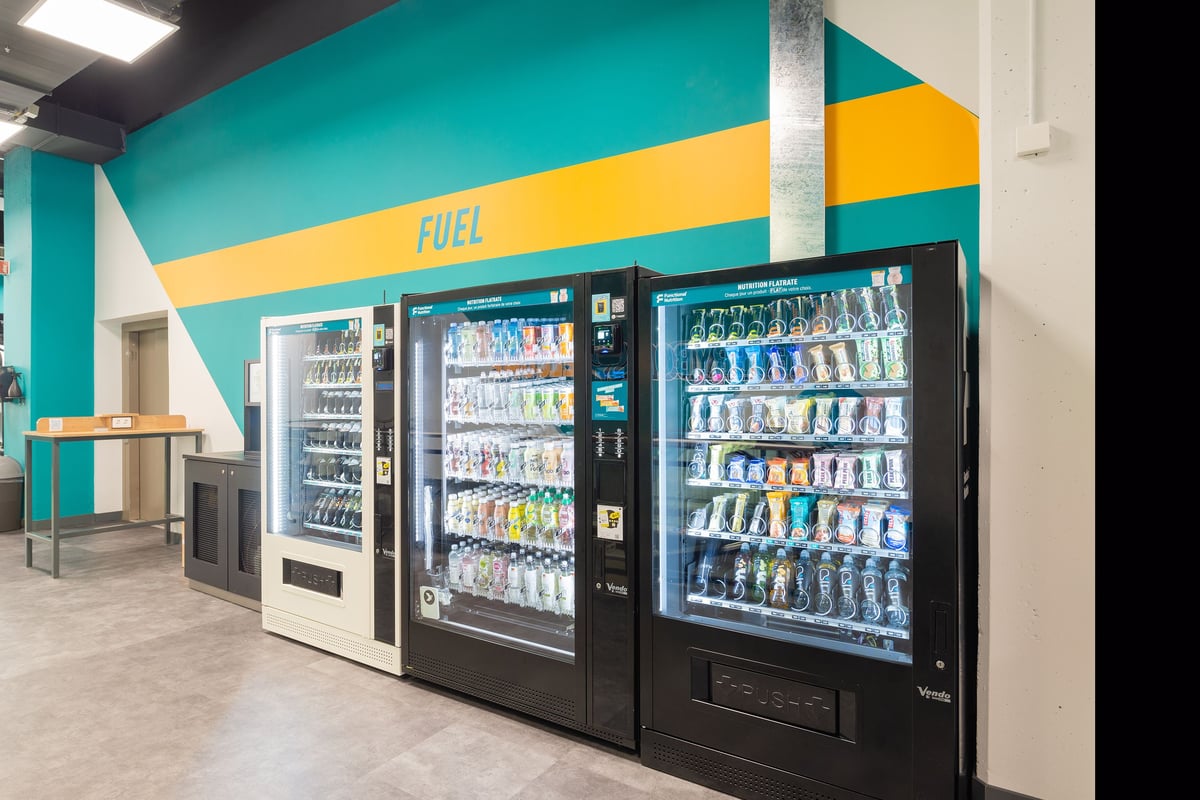 PureGym La Chaux-de-fonds: Vending machines filled with beverages and snacks in an open space with bright teal walls.