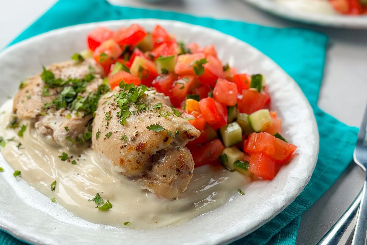 Grilled chicken with fresh herbs on white sauce beside chopped tomato and cucumber salad