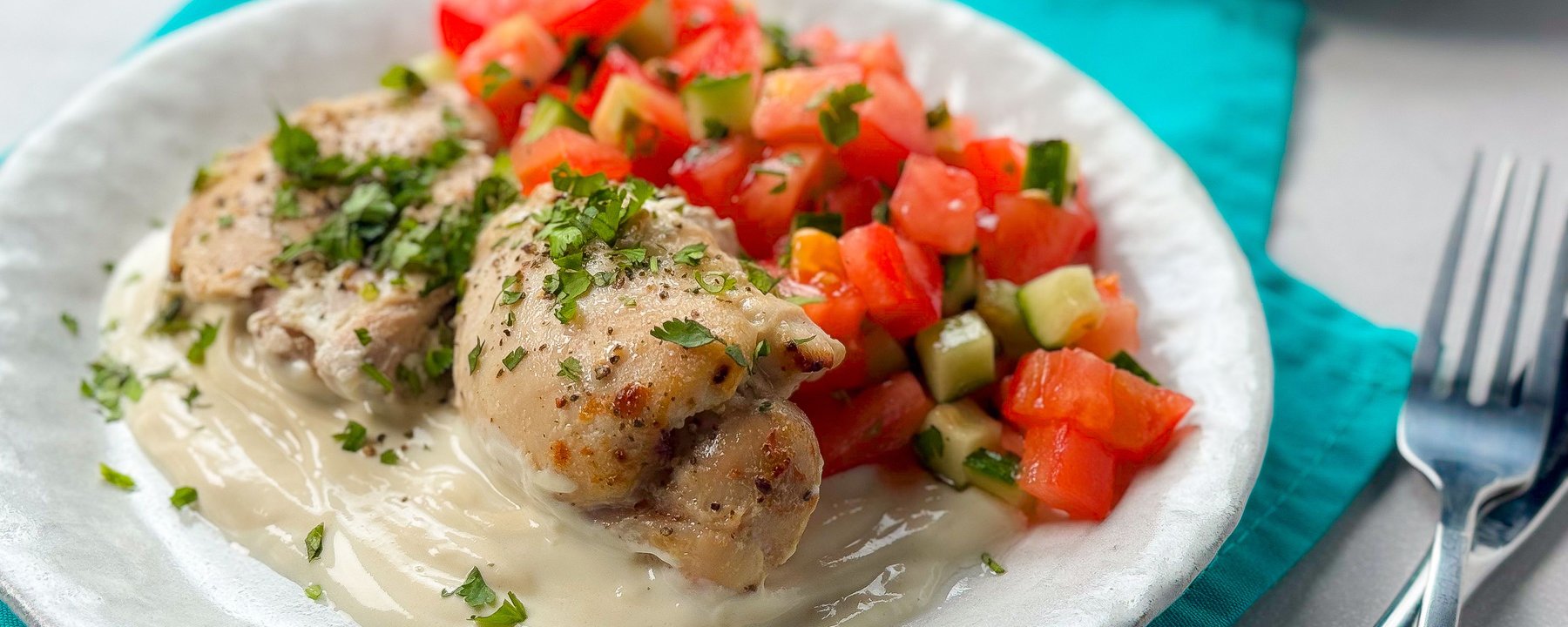 Grilled chicken with fresh herbs on white sauce beside chopped tomato and cucumber salad