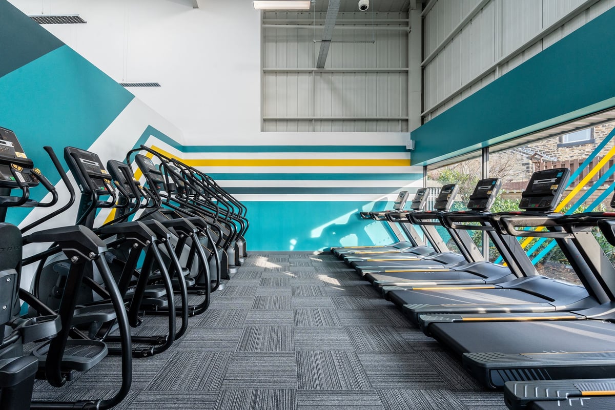 PureGym Keighley: Rows of treadmills in a colourful gym room with striped walls and mirrored panel.