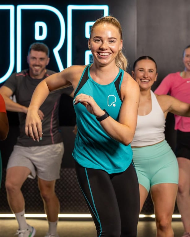 Donna sorridente in abbigliamento sportivo verde-blu che guida una sessione di fitness di gruppo energica con segnaletica al neon