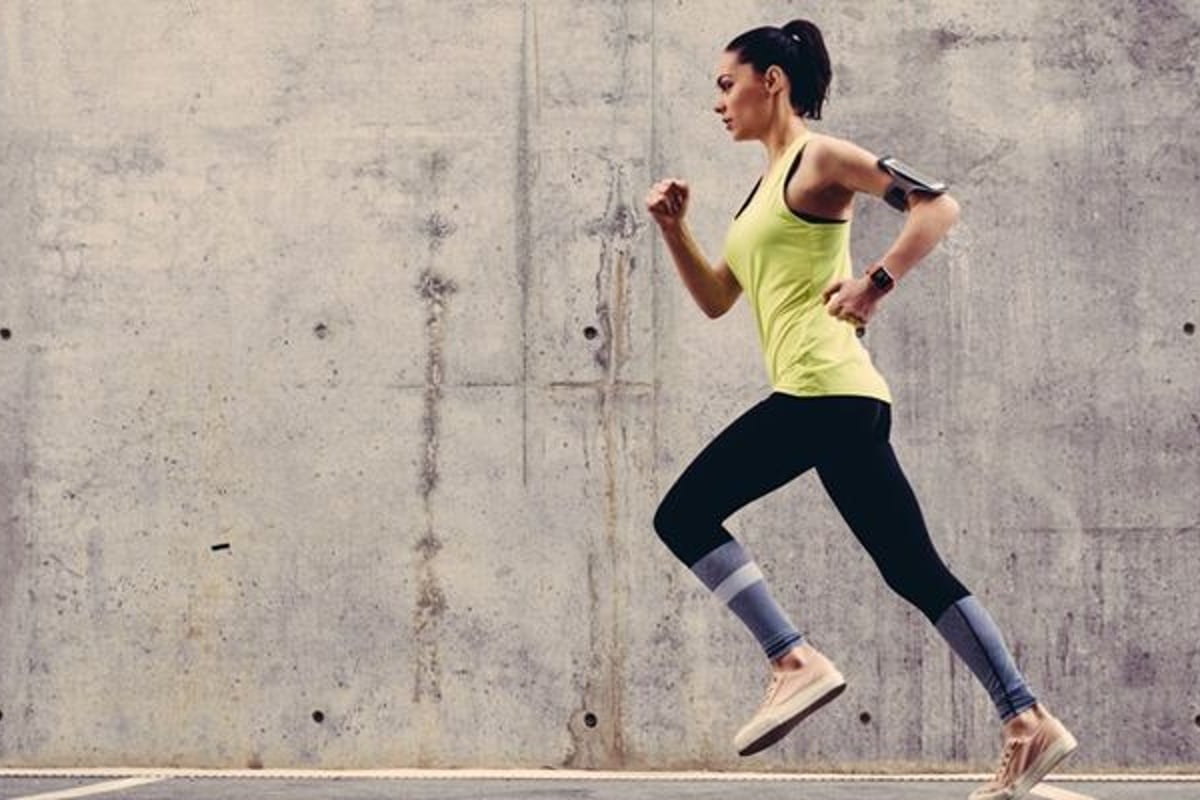 Woman in yellow vest and black leggings running outdoors against concrete wall