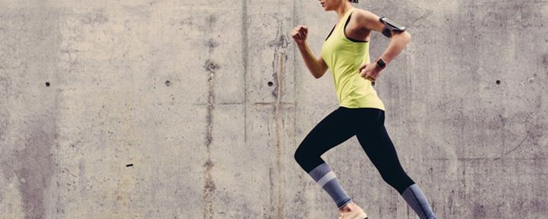 Woman in yellow vest and black leggings running outdoors against concrete wall