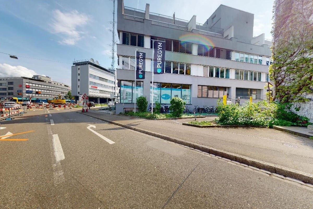 PureGym Basel Dreispitz: Modern city street with multistory buildings, storefronts, pedestrians, and parked bicycles.