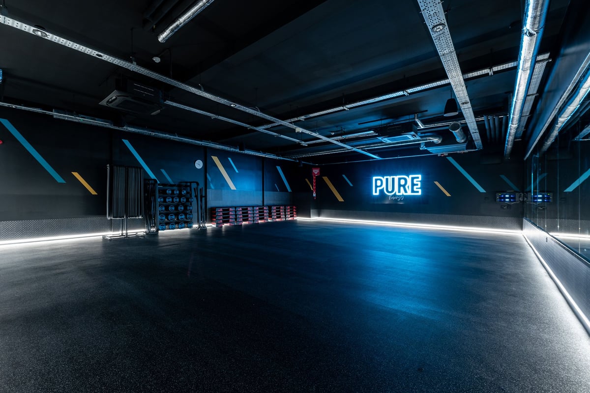 PureGym Ballymena: Large open studio space with blue lighting, bare walls, and rubberized flooring.