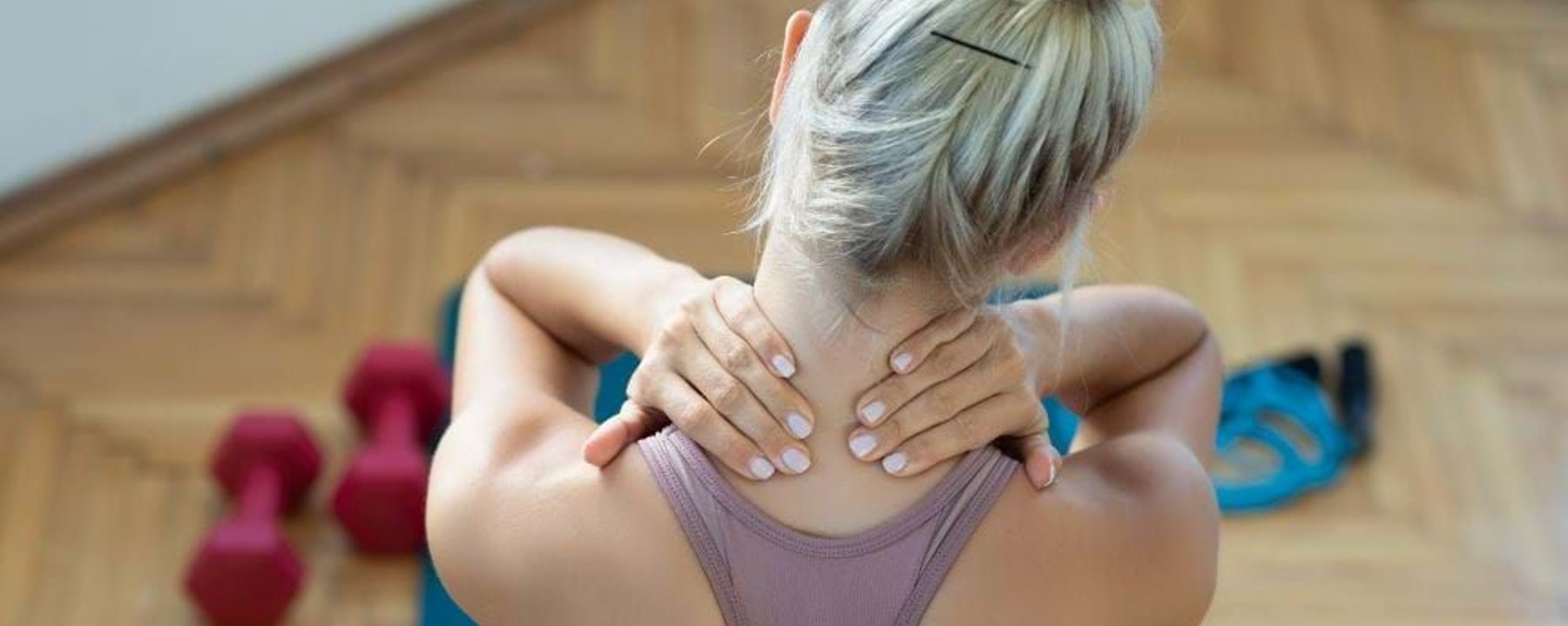 Woman with blonde hair in a bun holding her neck while sitting on a mat with dumbbells