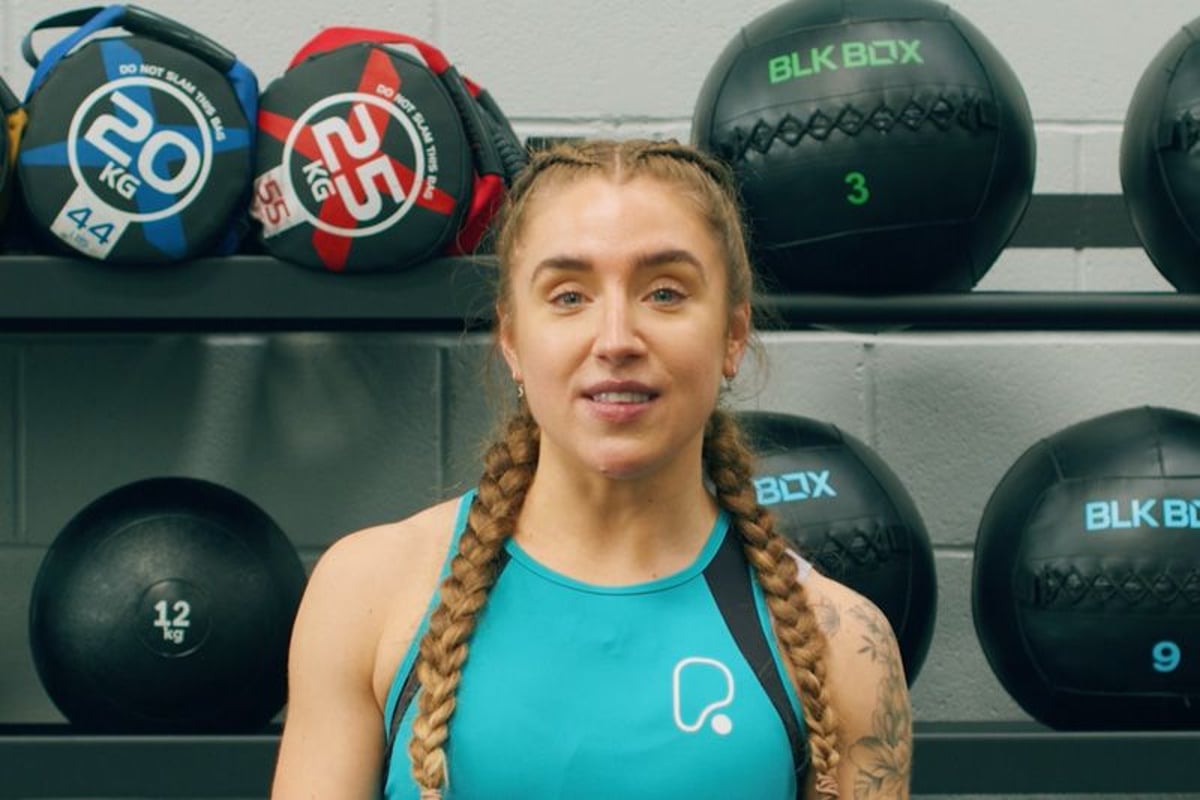 Woman with braided hair in turquoise sportswear standing in front of gym medicine balls on