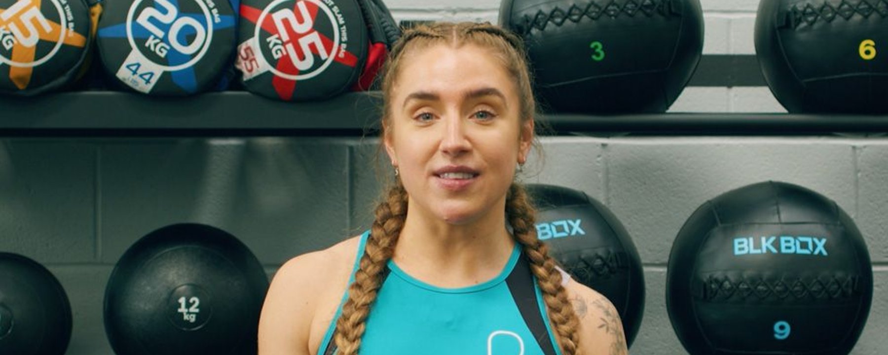 Woman with braided hair in turquoise sportswear standing in front of gym medicine balls on