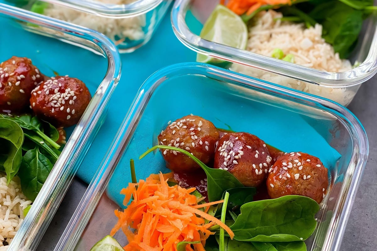 Meal prep containers with rice, shredded carrot, spinach, lime wedge, and meatballs with s
