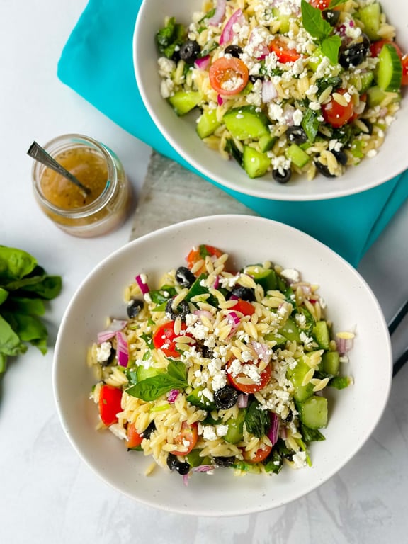 Two bowls of orzo salad with cherry tomatoes, cucumber, olives, feta, and herbs