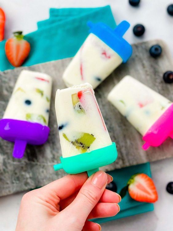 Hand holding a fruit-filled yoghurt popsicle with more on a stone board and scattered frui