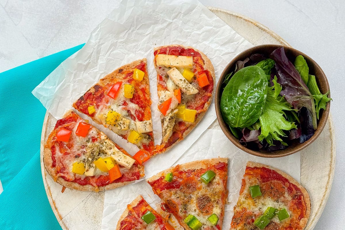 Two small pizzas with vegetables and a bowl of salad on a wooden board with a pizza cutter