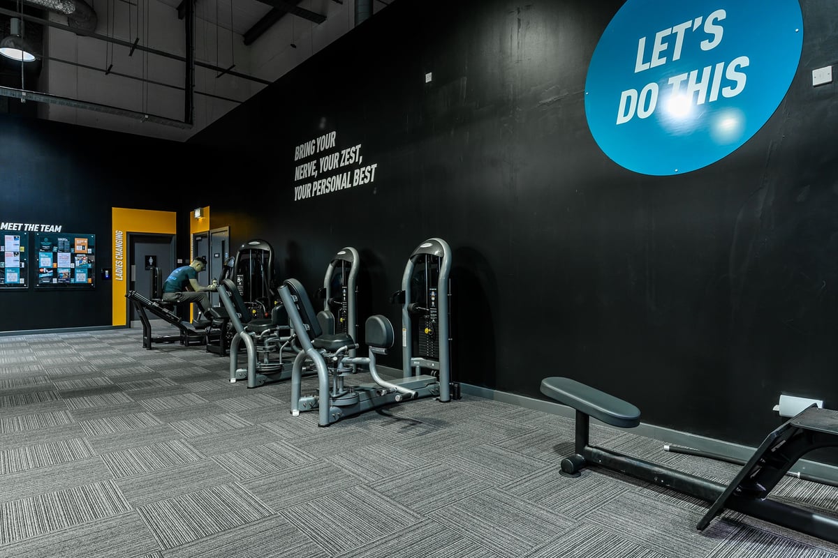 PureGym Bromborough: Weight machines, benches, and motivational signage in a dimly lit gym studio.