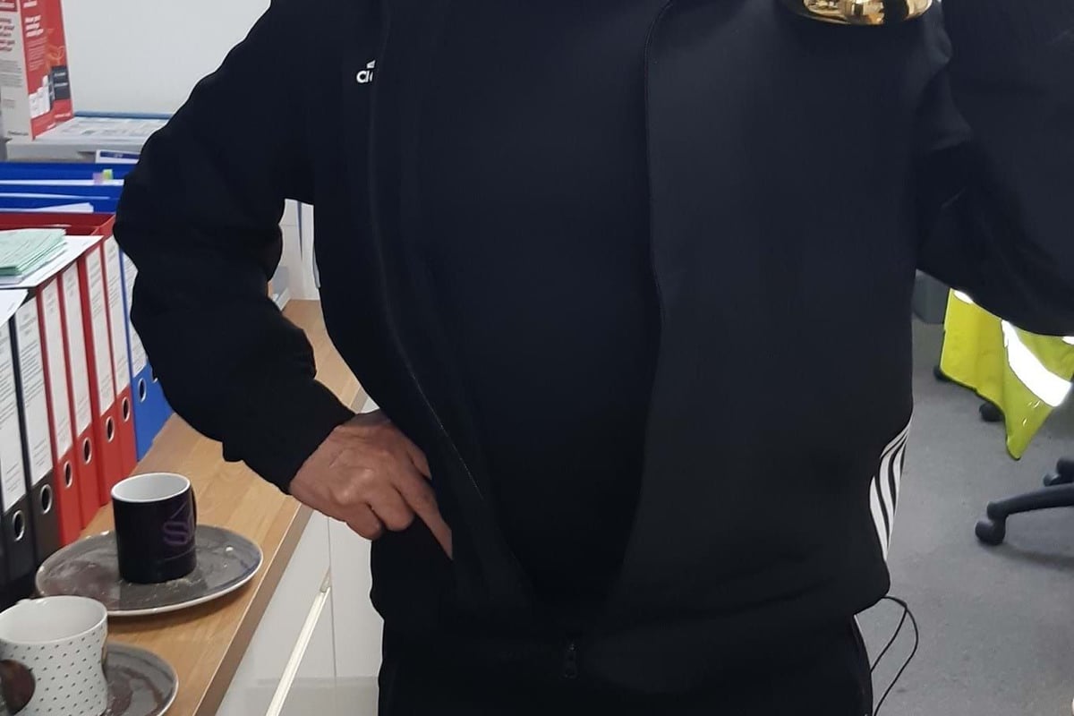 Man in black tracksuit holding a golden trophy in an office room with files and cups