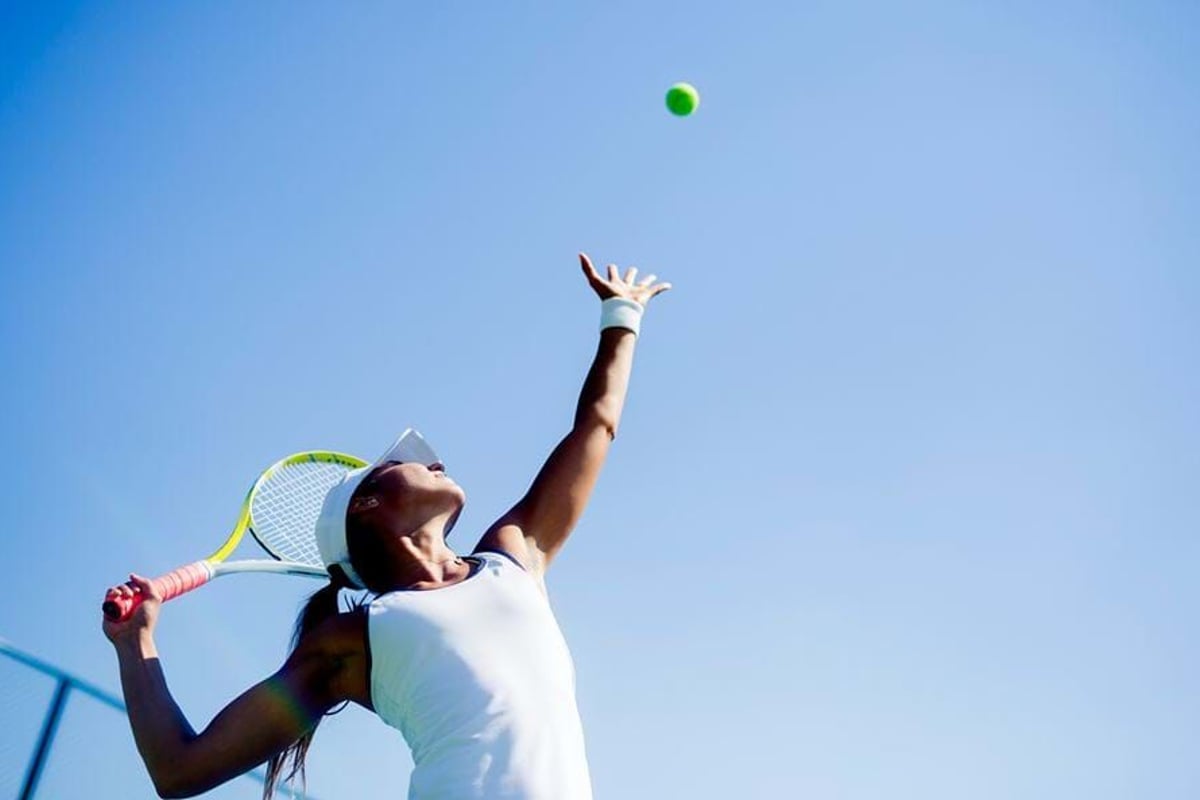 Female tennis player in white preparing to serve against clear blue sky