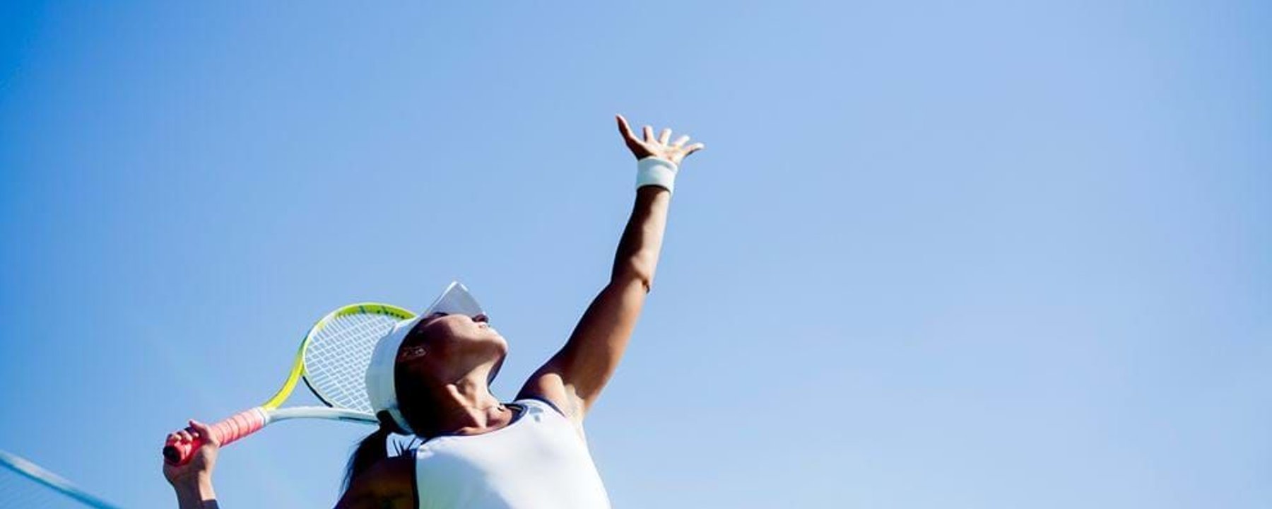 Female tennis player in white preparing to serve against clear blue sky