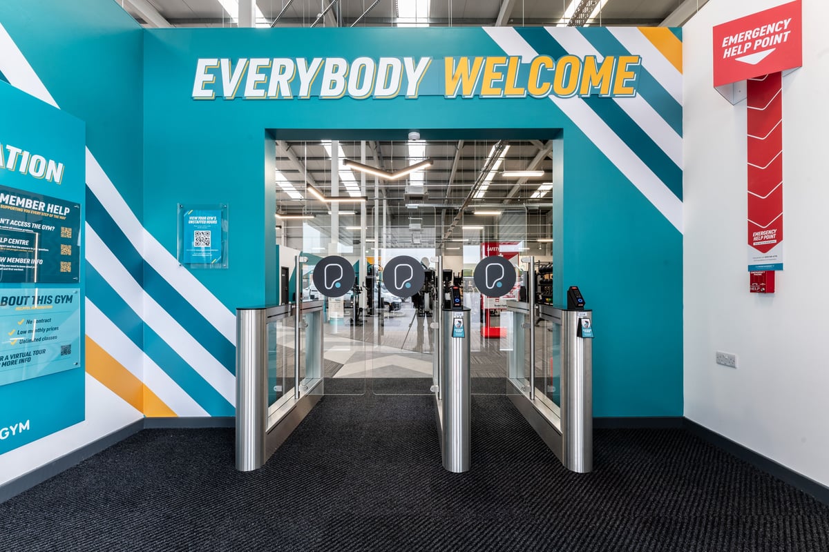 PureGym Port Talbot: Entry turnstiles, vibrant welcome signage, emergency equipment station.