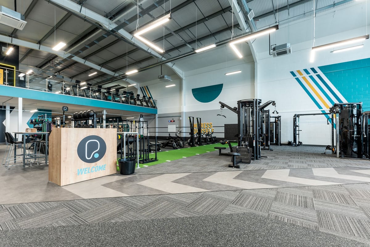 PureGym Liverpool Edge Lane: Functional training area with weight racks, benches, turf flooring, and front desk kiosk.