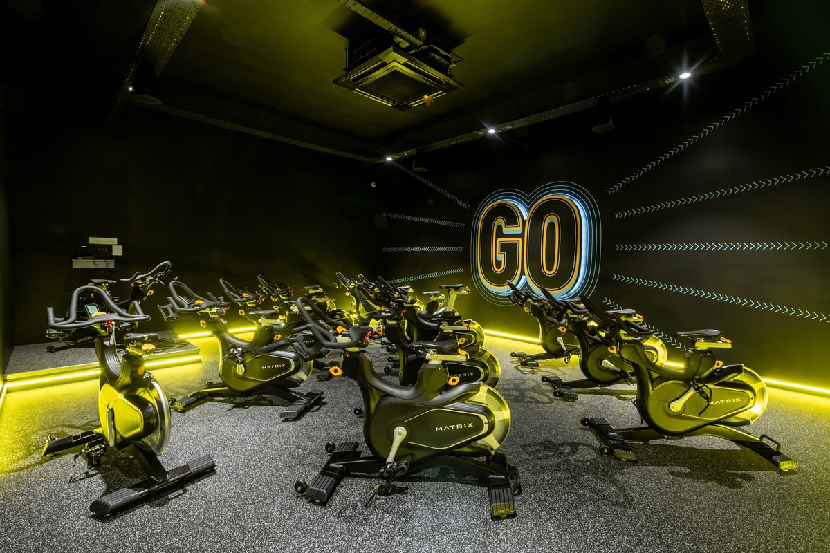PureGym Derby Kingsway Retail Park: Rows of stationary bikes in dimly lit studio emblazoned with neon 'GO' signage.