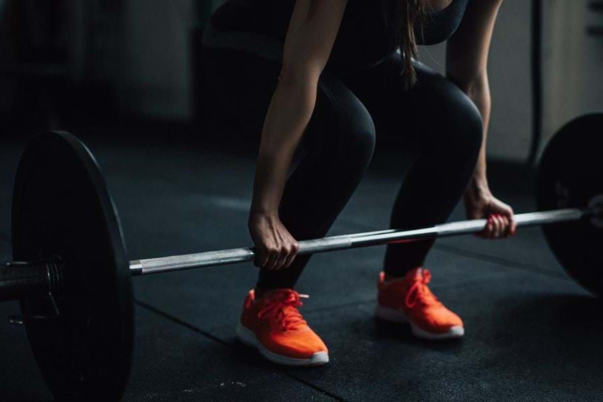 Person in black leggings and orange trainers lifting a barbell in a gym