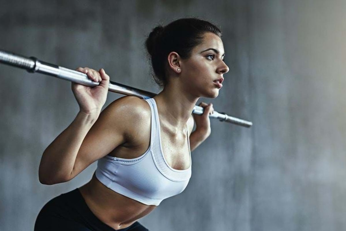 Woman in sports bra lifting a barbell on her shoulders against a grey wall