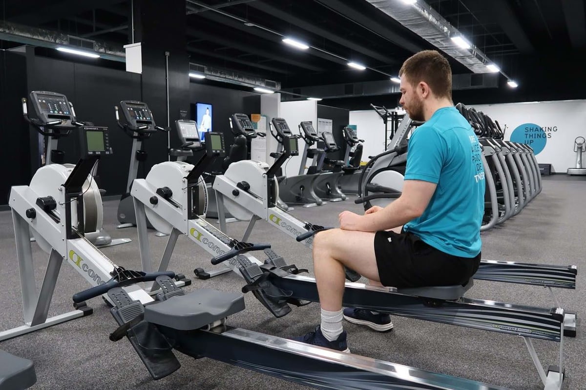 Man in blue shirt sitting on rowing machine in gym with exercise bikes in background