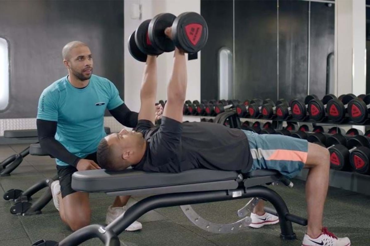 Man lifting dumbbells on bench press with trainer spotting in gym room.