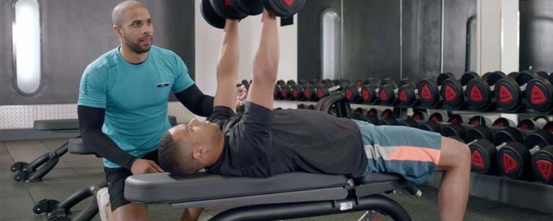Man lifting dumbbells on bench press with trainer spotting in gym room.