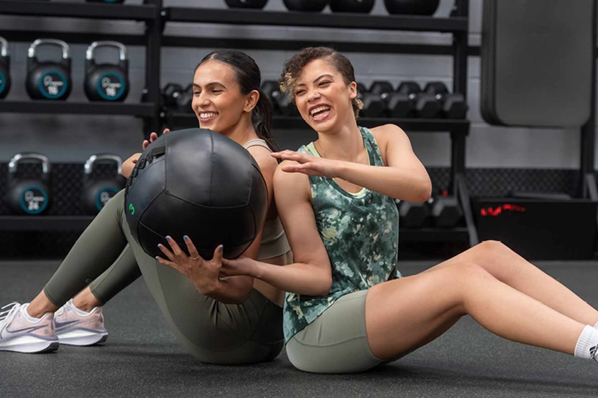 Zwei Frauen trainieren zusammen mit einem Medizinball im Fitnessstudio und lachen.