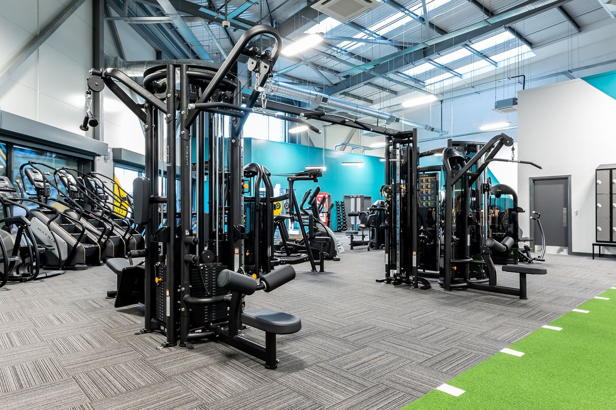 PureGym Kirkby: Strength training area with cable machines, squat racks, benches, and rubberized tiled flooring.