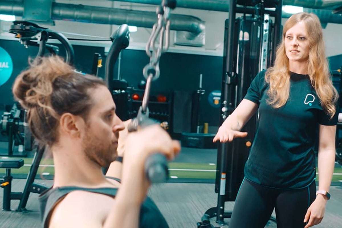 Man lifting weights while woman in sportswear coaches him in gym