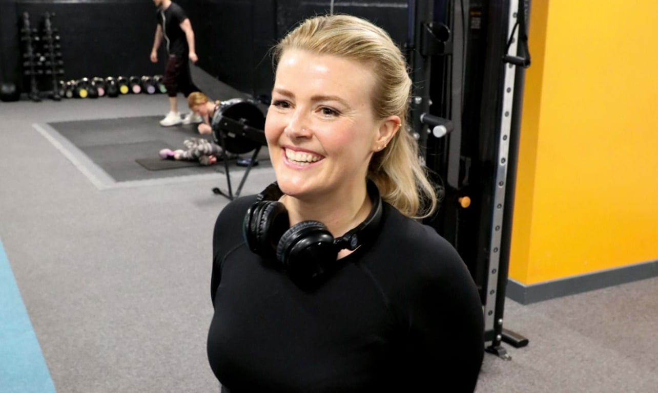 Smiling woman with headphones around her neck in a gym with people exercising behind her