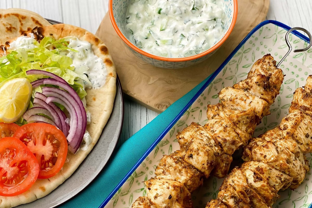 Grilled chicken skewers, flatbread with salad, and a bowl of tzatziki with lemon halves.