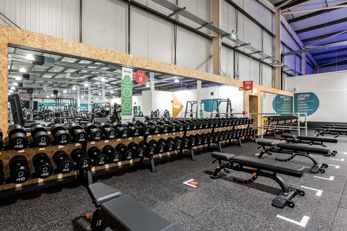 PureGym Swindon Stratton: Rows of weight benches, dumbbells, resistance machines, and gym signage on rubberized flooring.