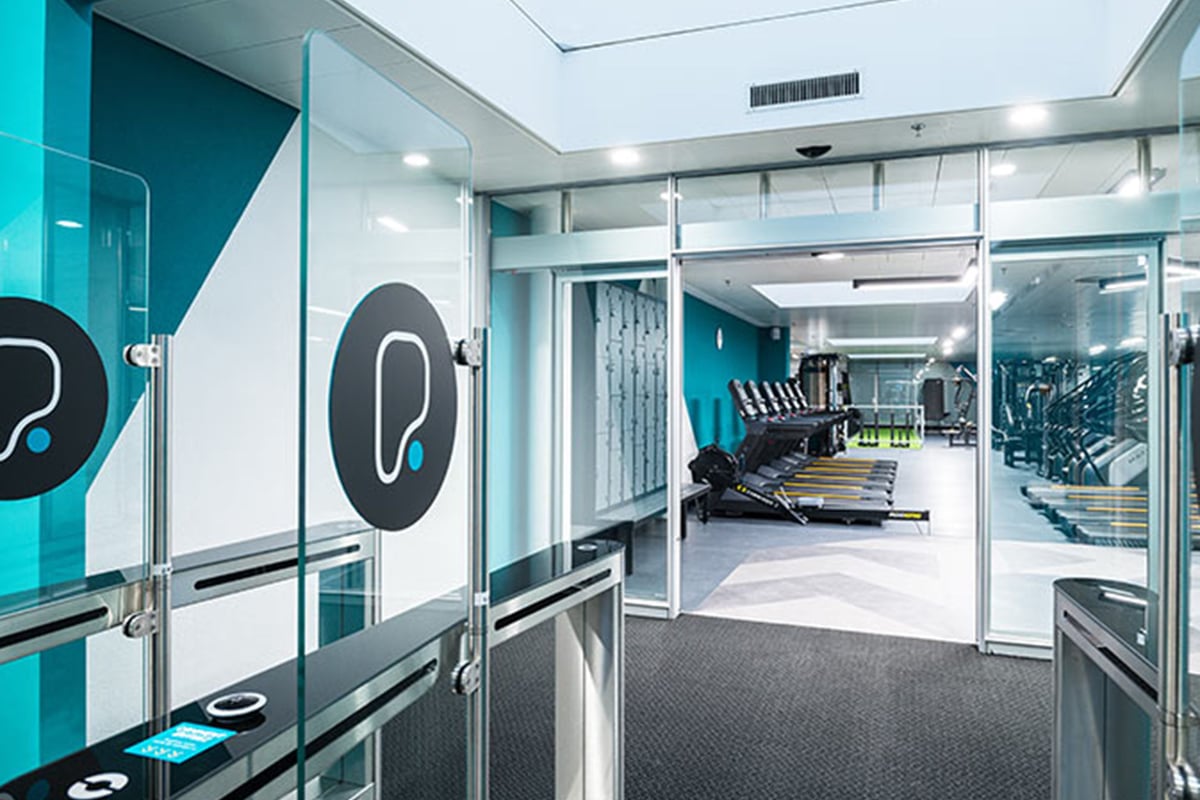 PureGym Fribourg: Entrée de la salle de sport avec cloisons vitrées, tapis de course visibles au-delà, accentués dans une palette de couleurs bleu sarcelle et noir.