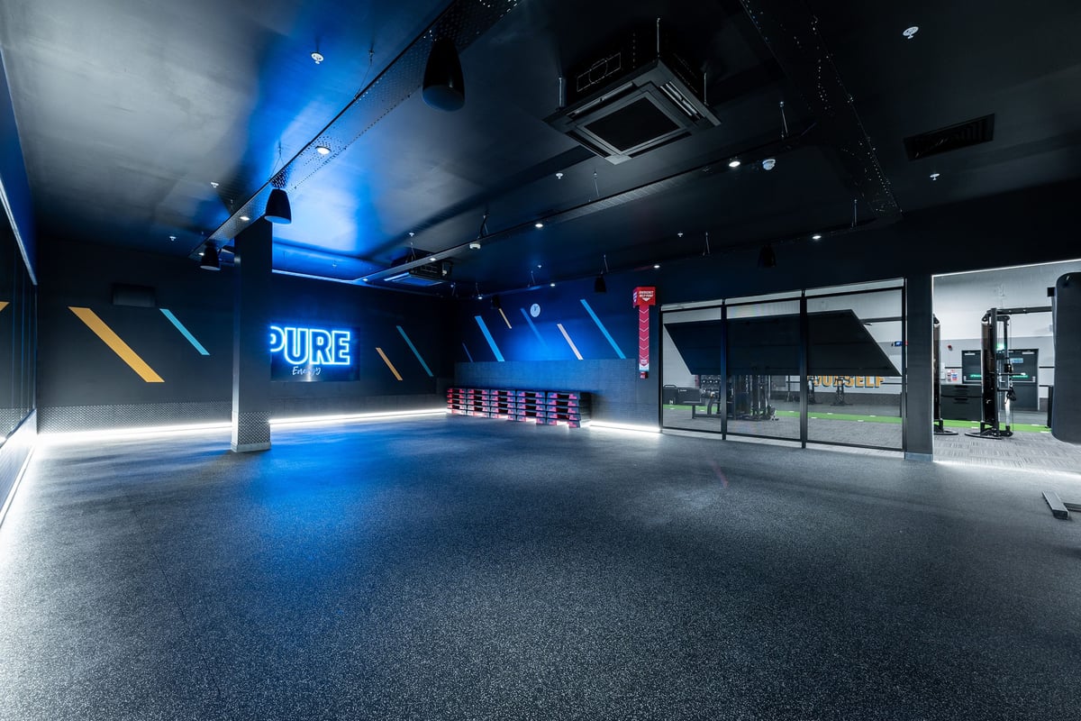 PureGym Rushden: Dimly lit studio with blue mood lighting, rubberized flooring, and workout rigs.