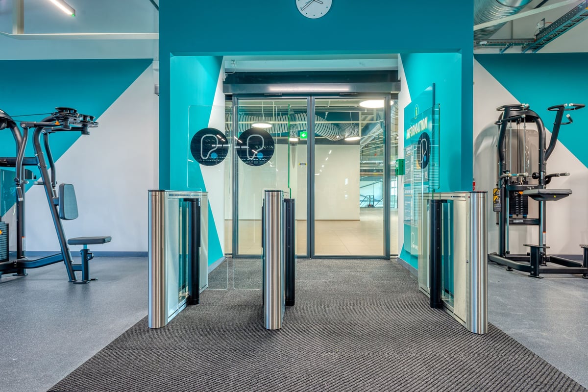 PureGym Martigny: Innenausstattung des Fitnessraums mit Kraftgeräten, Freihanteln, gläsernen Eingangstoren und blaugrünen Akzentwänden.