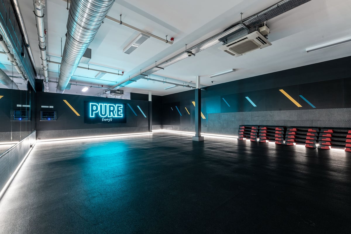 PureGym Ashton Under Lyne: Rows of red seats or benches, mirrored walls, teal and yellow lighting.