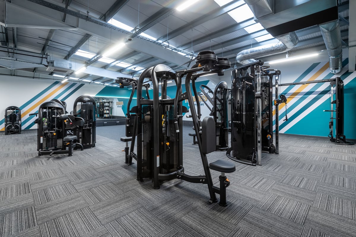 PureGym Derby Kingsway Retail Park: Weight machines, benches, rubberized flooring in a gym studio with teal accented walls.