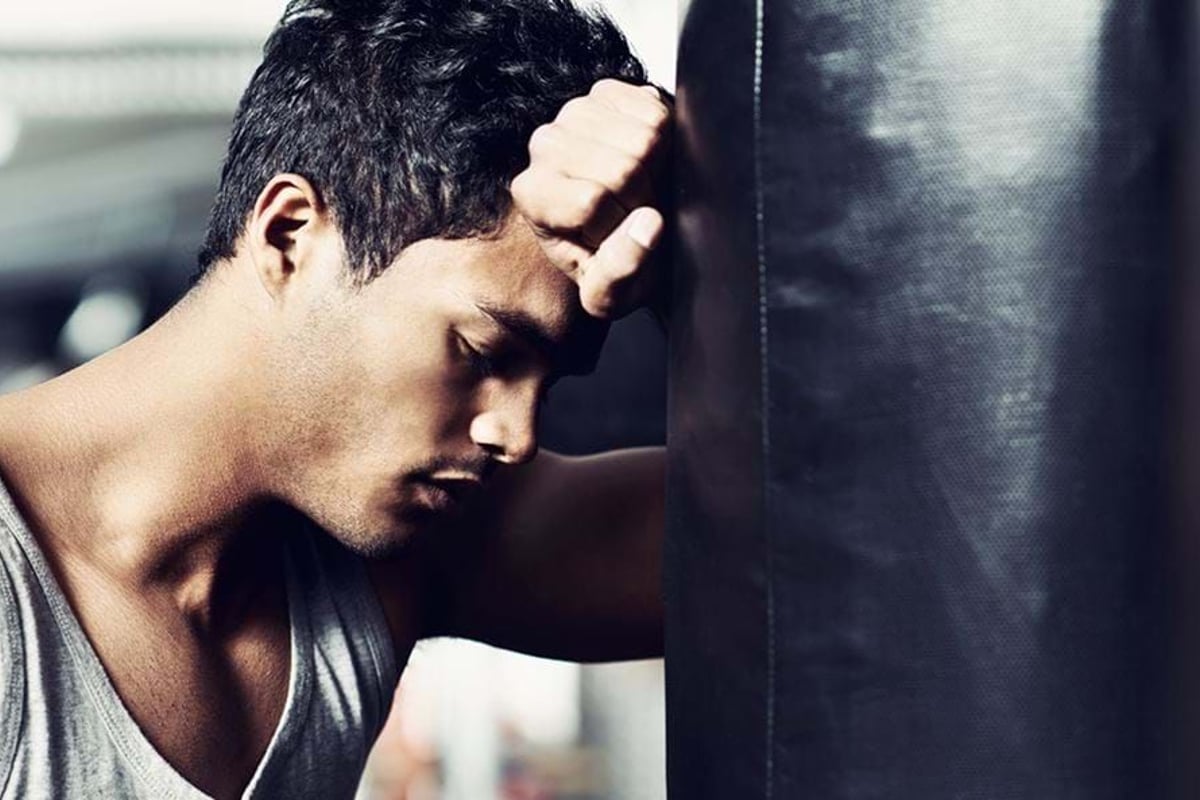 Man in grey tank top leaning forehead against a black punching bag with eyes closed