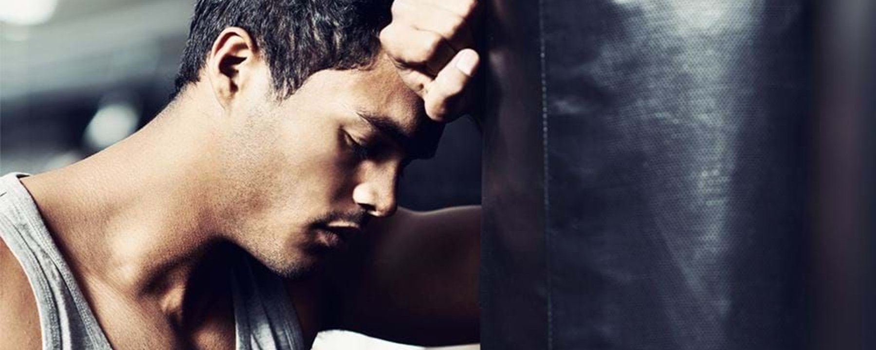 Man in grey tank top leaning forehead against a black punching bag with eyes closed