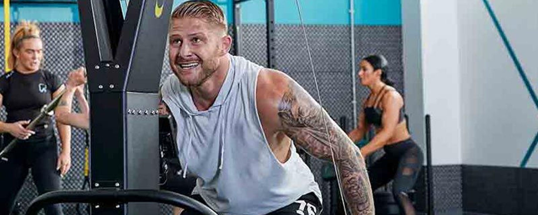 Homme au bras tatoué s’entraîne sur appareil de gym pendant que deux femmes font du sport