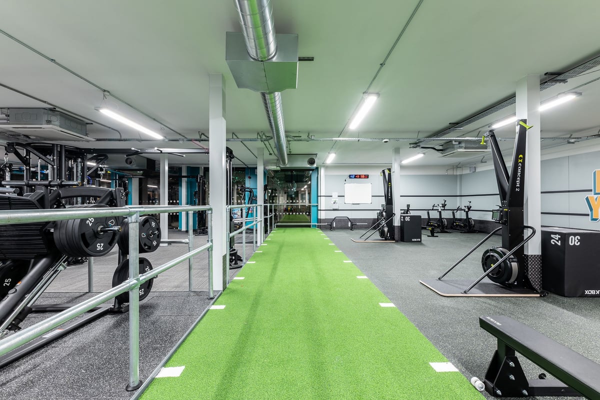 PureGym Manchester First Street: Green turf, weight racks, benches, mirrors, and rubberized flooring inside a gym studio.