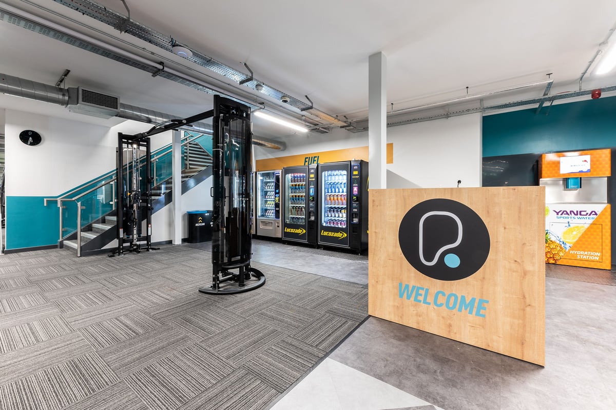PureGym Manchester First Street: Weight machines, vending units, cushioned flooring, welcoming sign with logo.