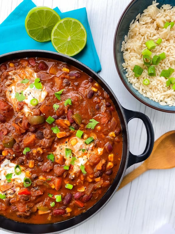 Chilli con carne with sour cream and herbs in a pan next to a bowl of rice and lime halves