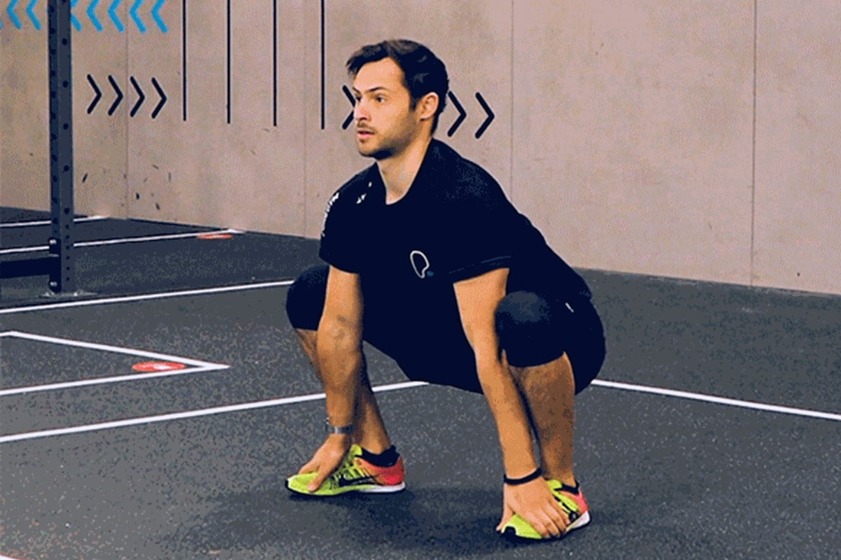 Man in black sportswear performing deep squat on gym floor with yellow trainers